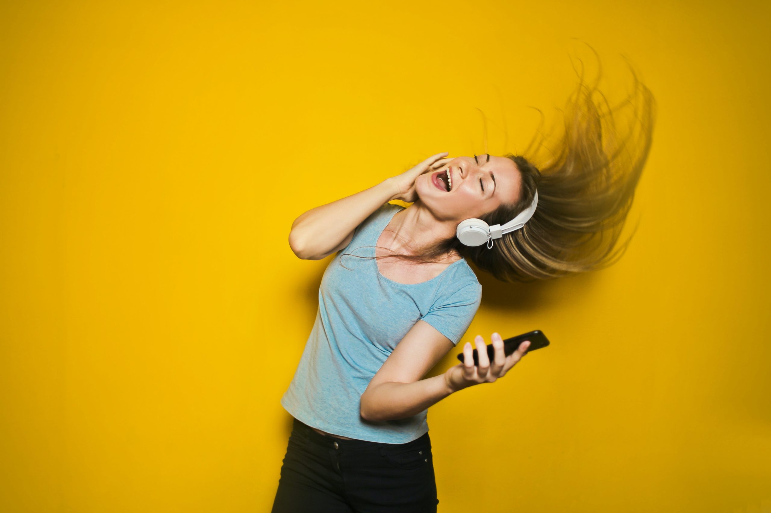 Eine junge Frau tanzt mit geschlossenen Augen und Kopfhörern auf den Ohren vor einer gelben Wand und hält ein Smartphone in der Hand.